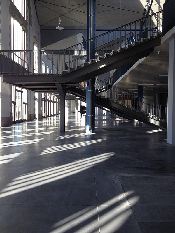 A wide architectural interior — concrete columns and a tall window casting a long shadow on a polished concrete floor.